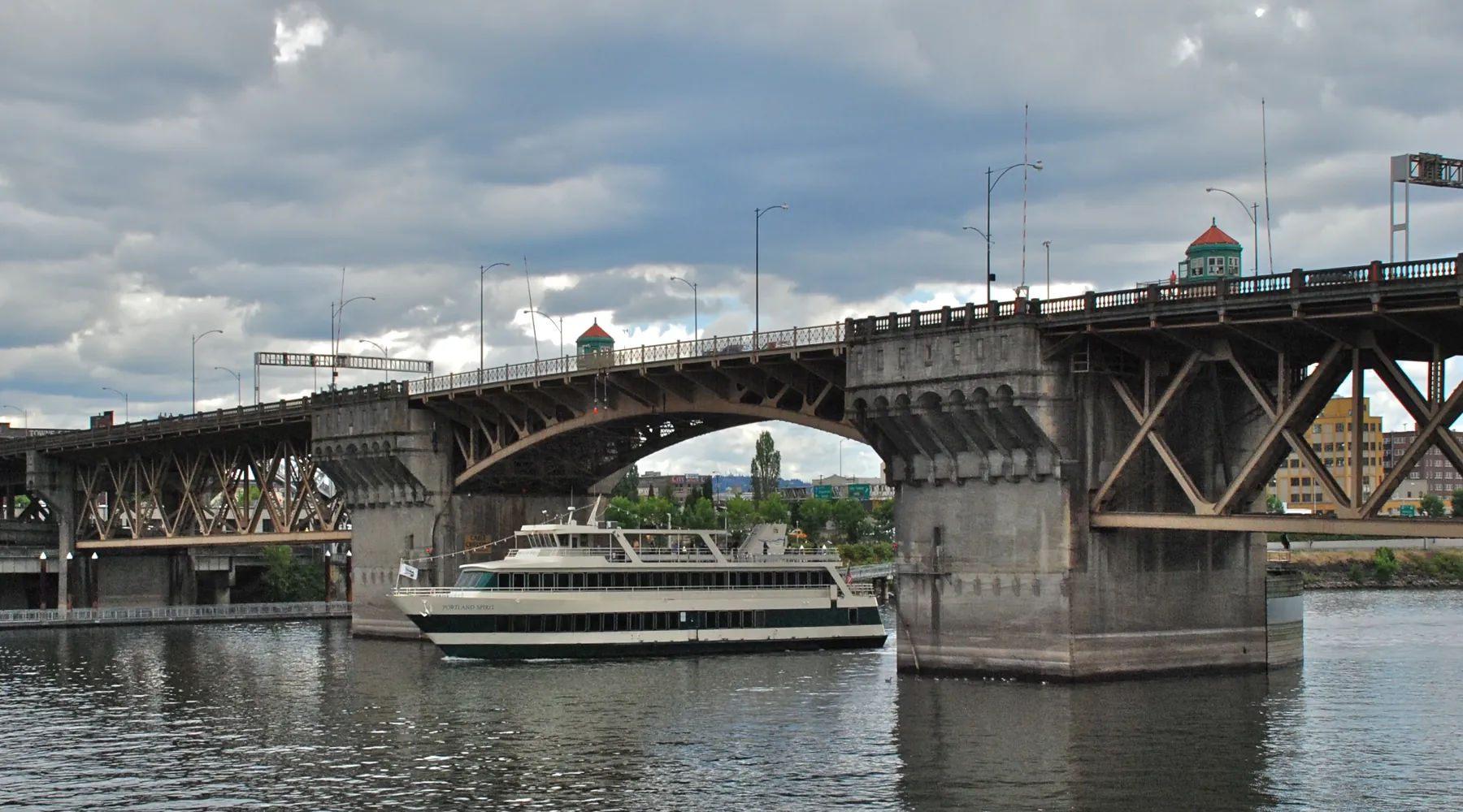 A cruise boat passing under the Burnside Bridge, with the concrete piers and steel arches above a grey-green river