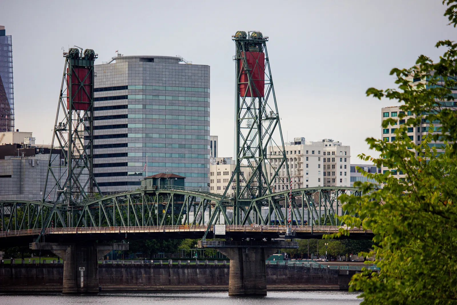 The two green lift towers of the Hawthorne Bridge with their red counterweights, rising behind a downtown office tower