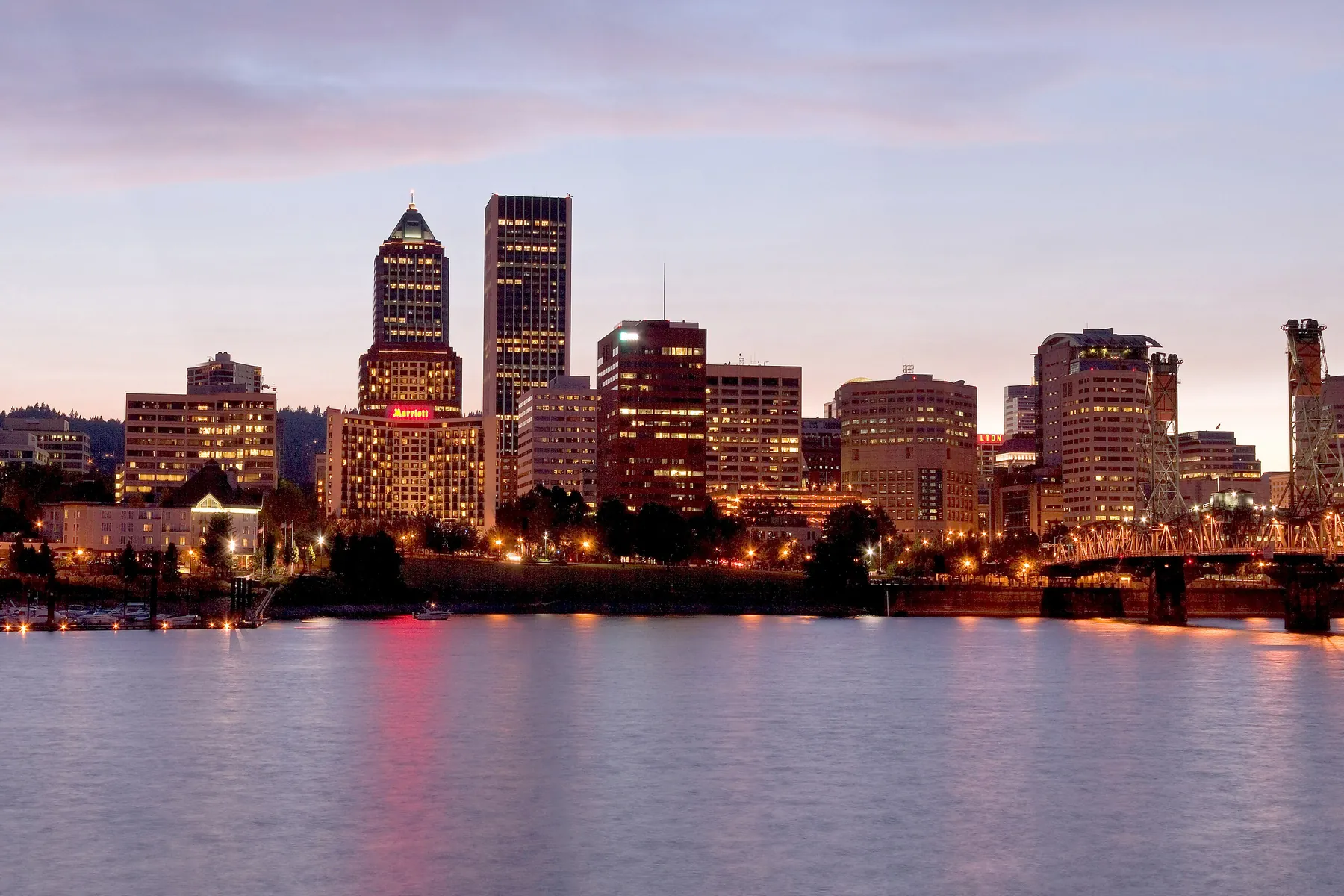 Downtown Portland towers glowing at dusk above the calm Willamette River, with warm lights along the bank