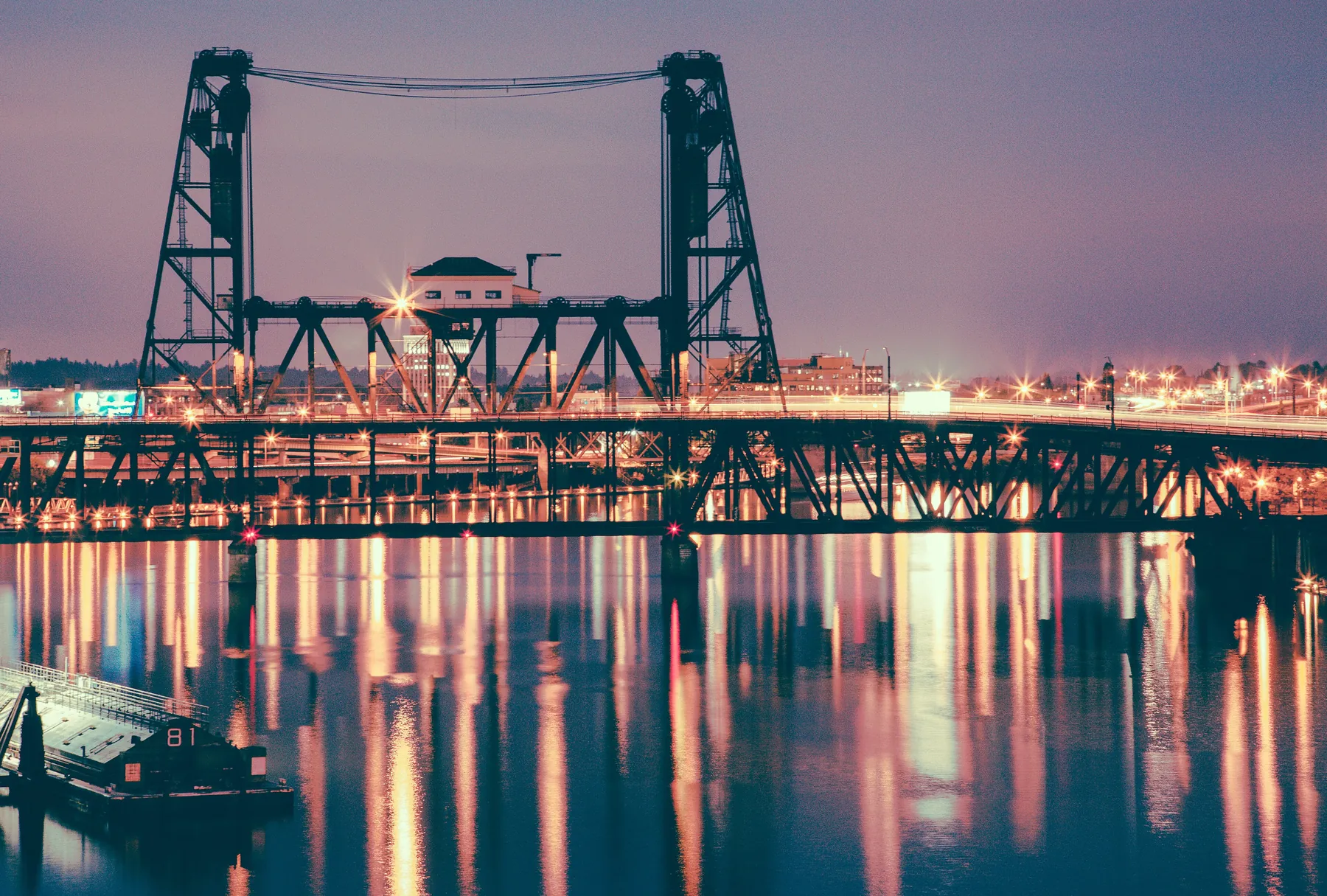 The Steel Bridge's twin lift towers lit at night, with city lights reflected in long streaks on the river
